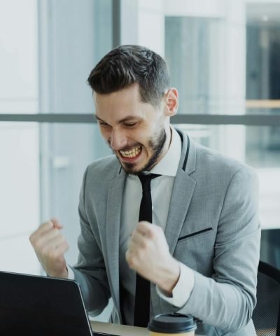 Man in grey suit celebrating success at his desk with a laptop.