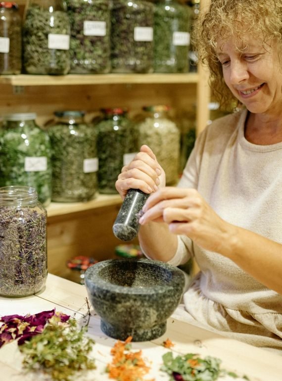 Woman grinding herbs with mortar and pestle in a cozy workshop filled with jars and dried flowers.